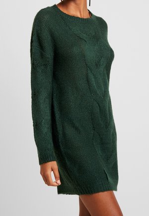 Ciemnozielona sukienka sweterkowa z długim rękawem, wykonana na drutach z wzorem warkoczy, noszona przez osobę na neutralnym tle.