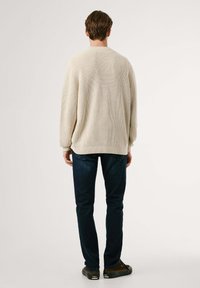 Beige gestrickter Pullover mit gerippter Textur, lockerer Passform und langen Ärmeln, kombiniert mit dunkelblauben Skinny Jeans und braunen Schuhen.