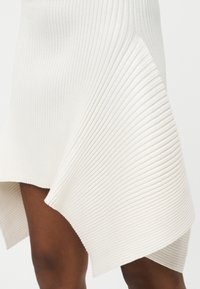 Λευκή ριγέ ασύμμετρη φούστα που πέφτει πάνω σε καστανά πόδια, με φωτεινό απλό φόντο.