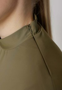 Olijfgroene top met een hoge kraag en een ritssluiting aan de zijkant. Stof lijkt glad en rekbaar met een matte afwerking. Close-up van de halslijn.