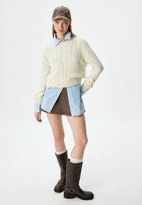 Creme kabelgebreide trui, lichtblauwe overhemddas eronder, bruin houndstooth rok, kniehoge bruine laarzen met creme sokken, bruine leren pet.
