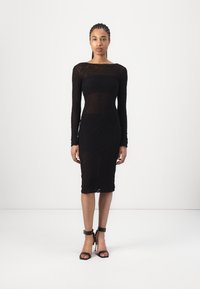 Zwart, lang mouwen jurk met een getailleerde silhouet en kanttextuur. Heeft een bootnek en een knie-lengte zoom. Gemaakt in combinatie met zwarte sandalen met bandjes.