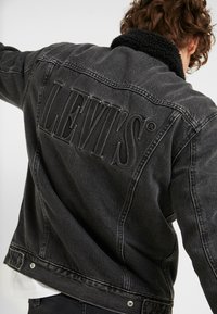 Czarna dżinsowa kurtka z teksturowanym czarnym kołnierzem z sztucznego futra. Posiada wytłoczone logo "LEVI'S" na plecach oraz typowe okucia kurtki dżinsowej.