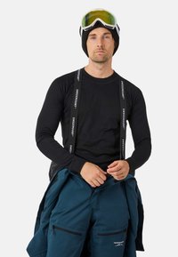 Zwarte geribbelde lange mouwen thermoshirt met verstelbare bretels; model draagt een beanie en skibril, gecombineerd met teal sneeuwbroeken.