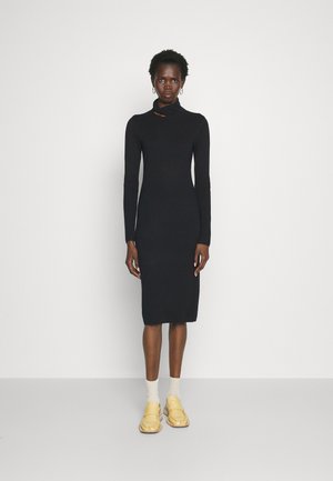 Μαύρο φόρεμα με μακριά μανίκια και ψηλό γιακά, με κοπή λεπτομέρειας, σφιχτή σιλουέτα, φτιαγμένο από μαλακό ύφασμα. Συνδυασμένο με κίτρινες loafers και υφασμάτινες κάλτσες.