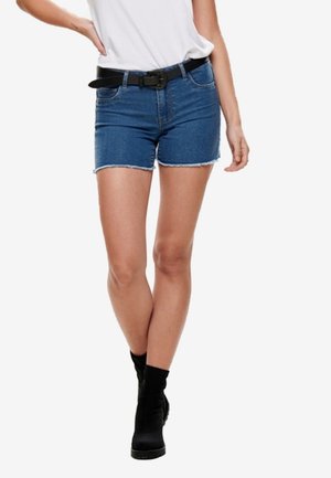 Vrouw draagt blauwe denimshorts met rafelige zoom, een wit ingestopt topje, een zwarte riem en zwarte enkellaarzen, staand met gekruiste benen.