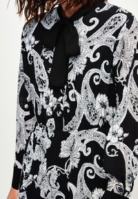 Vestido de estampado paisley en blanco y negro con un cuello curvado, detalle de lazo y tejido texturizado. Presenta acentos florales y cintura fruncida.