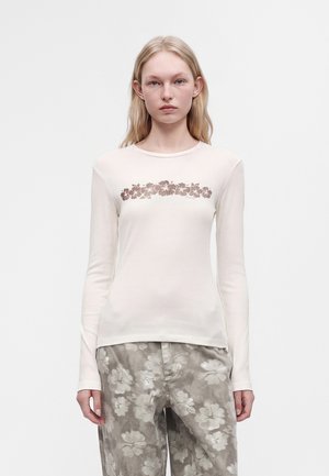 Jeune femme portant une chemise blanche à manches longues avec un motif floral marron et un pantalon ample gris avec des motifs floraux blancs.