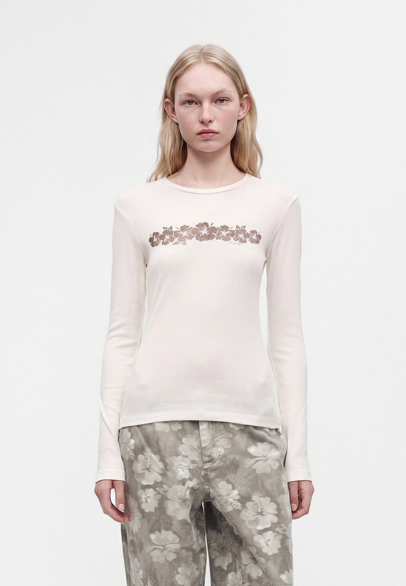 Jeune femme portant une chemise blanche à manches longues avec un motif floral marron et un pantalon ample gris avec des motifs floraux blancs.