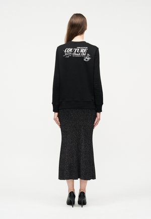 Ženska z dolgimi rjavimi lasmi nosi črno majico s kapuco z belim napisom "Couture Beach Club" in temno sijočo midi krilo ter črne pete, pogled od zadaj.