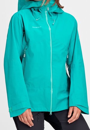 Chaqueta impermeable azul verdoso con cremallera frontal, capucha ajustable, bolsillos laterales con cremallera y tiras de velcro en las muñecas, usada por una persona con pantalones negros.