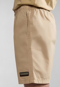 Beige shorts med elastisk talje, sidelomme og et sort og gult logo-patch nær kanten. Glat, letvægtigt stof.
