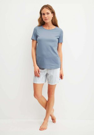 Donna in piedi a piedi nudi che indossa una maglietta blu a maniche corte e pantaloncini rigati azzurri e bianchi su uno sfondo bianco semplice.