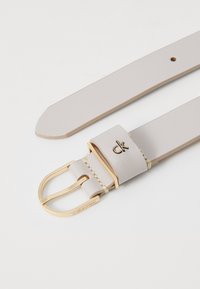 Hellgrauer Leder-Gürtel mit glatter Textur, ausgestattet mit einer goldenen Metallschnalle und einem kleinen, in das Band nahe der Schnalle geprägten Logo.