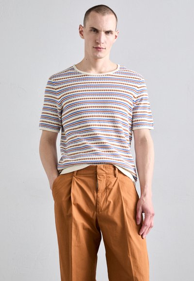 T-shirt à rayures à manches courtes dans les couleurs bleue, orange et blanche, avec un col rond. Associe à un pantalon ample orange-brun.