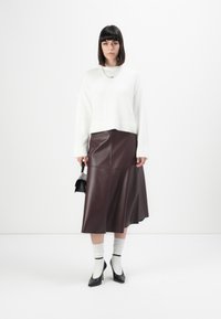 Vrouw in een witte fluffy trui, een bruin leren midi-rok, witte sokken met zwarte hakken, met een kleine zwarte handtas, staande tegen een witte achtergrond.