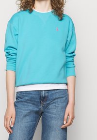 Světle modrá mikina vyrobená z měkkého materiálu, s kulatým výstřihem, žebrovanými manžetami a malým růžovým logem na levé hrudi. Nositá přes bílou košili.