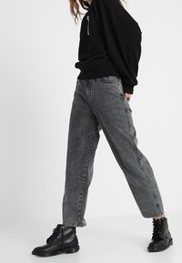 Felpa nera, jeans grigi in stile loose-fit e stivali neri stringati. I jeans hanno tasche laterali e un lavaggio leggermente sbiadito per un aspetto texturizzato.
