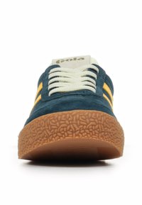 Zapatilla de ante azul con rayas amarillas, suela de goma marrón texturizada y cordones crema. Logo de la marca visible en la lengüeta.