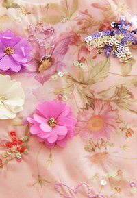 Tela floral en suave rosa con flores de papel superpuestas en lavanda, blanco y rosa, adornadas con lentejuelas y acentos bordados.
