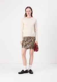 Krems krāsas džemperis, leopardu raksta mini svārki, melni mokasīni, potītes zeķes, tur rokās mazu sarkanu somiņu. Gluda tekstūra, pieguļošs dizains.