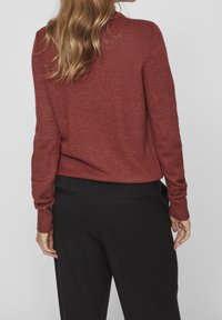 Femme aux cheveux châtain clair ondulés portant un pull texturé couleur rouille et un pantalon noir sur mesure, debout devant un fond gris uni.