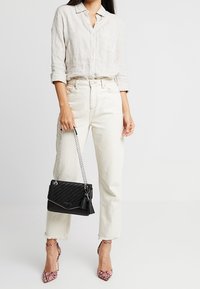 Borsa a tracolla nera trapuntata con catena argentata, con un accentuato pon-pon e dettagli borchiati. La modella indossa una camicia di lino chiaro, jeans bianco sporco e tacchi rosa.