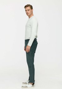 Férfi világos pulóverben és sötét kordnadrágban áll oldalt egy sima fehér háttér előtt, fehér és szürke tornacipőt viselve.