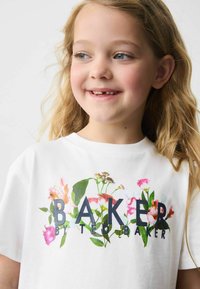Valge puuvillane T-särk, millel on lilleline kujundus roosa ja rohelise aktsendiga, kus on kujutatud tekst "BAKER BY TED BAKER" julgete tumedate tähtedega.