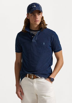 Marineblauwe polo met een gestructureerde afwerking, korte mouwen en een kraagdetail. Draagt over een gestreept shirt, gecombineerd met witte broek en een bruine riem.
