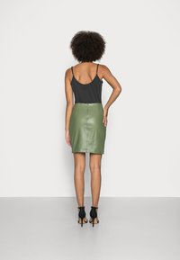 Donna con capelli ricci che indossa un top senza maniche nero, una gonna di pelle verde oliva e sandali con tacco alti neri, in piedi contro uno sfondo bianco.