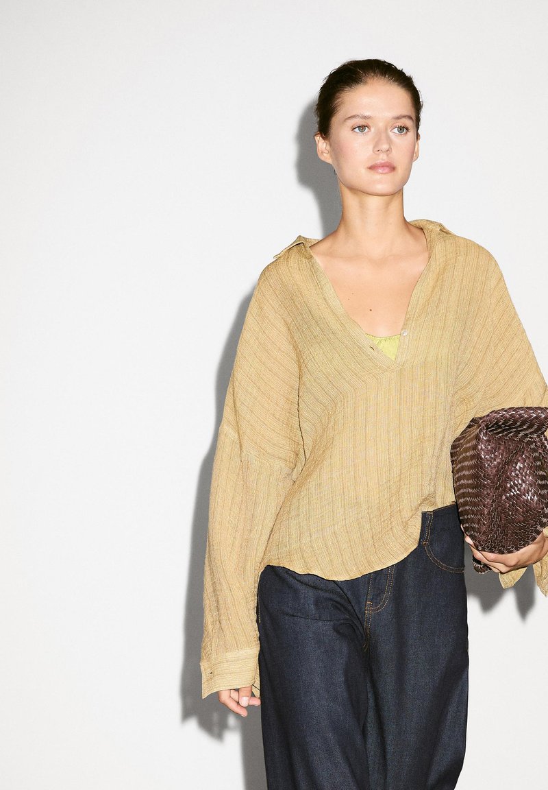 Světle žlutá oversized košile s texturovanou tkaninou, výstřihem do V a spadlými rameny, spárovaná s tmavě modrými širokými džínami a tkanou kabelkou.