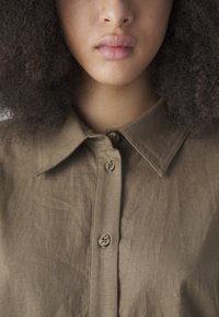 Camicia marrone con colletto in un tessuto testurizzato, con bottoni e dettagli di cucitura visibili. Il tessuto appare morbido e leggermente stropicciato.