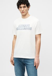 Νεαρός άνδρας με σγουρά μαλλιά, που φοράει λευκό t-shirt Armani Exchange και μπλε τζιν, στέκεται μπροστά σε ένα απλό φωτεινό φόντο.