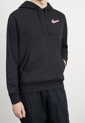 Personne portant un sweat à capuche noir avec poche avant et logo Nike Swoosh rose, mains partiellement dans les poches, sur un fond blanc uni.
