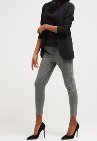 Zwarte blazer over een zwarte top, gecombineerd met grijskleurige nep leren broek met ritsen. Zwarte puntige hakken maken de look compleet.