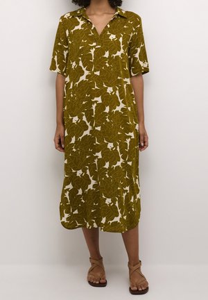 Kvinde iført en knælang, kortærmet kjole med olivengrønt bladprint og åben krave, parret med beige sandaler med remme.