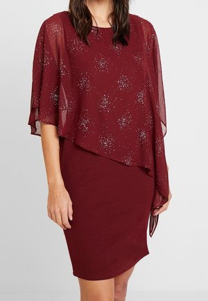 Femme portant une robe bordeaux sans manches avec une surcouche asymétrique transparente ornée de motifs argentés scintillants.