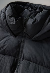 Fekete pufi dzseki magas gallérral és cipzáras zárral. Texturált, könnyű anyagból készült, vízszintes varrásrészletekkel.