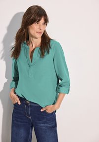 Camicia turchese a maniche lunghe con scollatura a V e polsini arrotolati, abbinata a jeans scuri in denim, dotati di tasche anteriori e vestibilità rilassata.