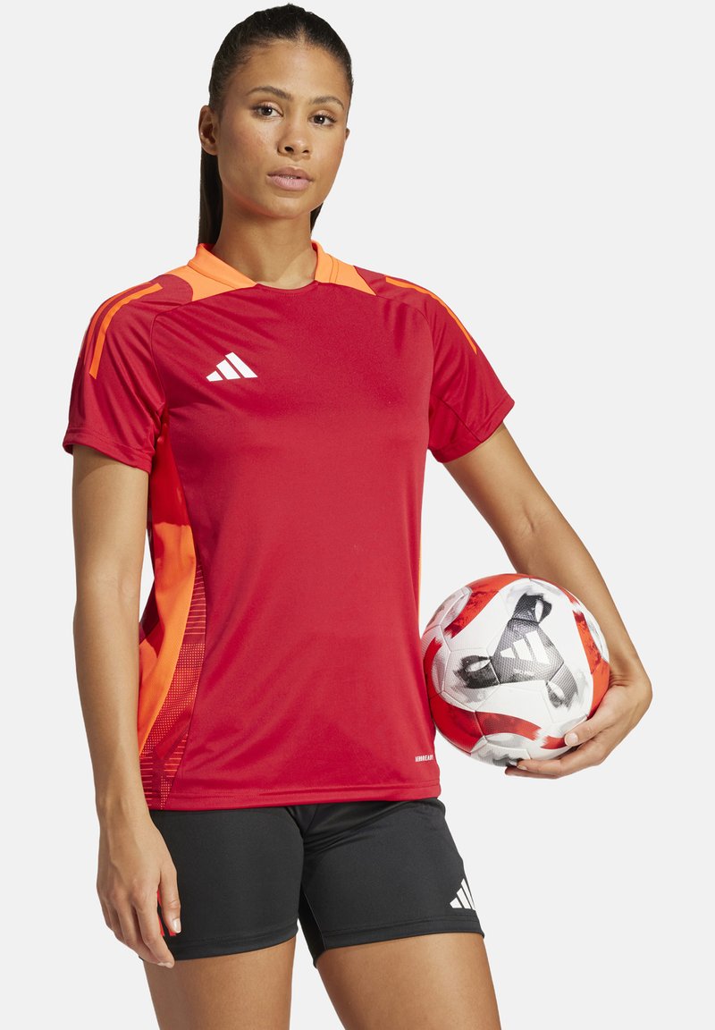 Maglia sportiva rossa con accenti arancioni e tre strisce, abbinata a pantaloni corti neri. Il giocatore tiene in mano un pallone da calcio a motivo.