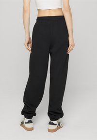 Moteris dėvinti laisvus juodus sportinius kelnes ir baltus sportinius batelius, stovinti nugara į kamerą priešais paprastą foną.