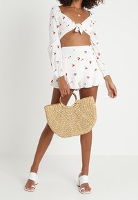Valkoinen crop-top, jossa on kirsikkakuosi ja nauhakiinnitys edessä, yhdessä sopivan flare-mallisessa mini-hameessa. Malli pitää pyöreää olkakassia ja käyttää valkoisia korkokenkiä.