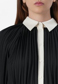 Close-up of a woman’s top featuring black pleated fabric, a white collar, and button details, emphasizing the stylish design elements.