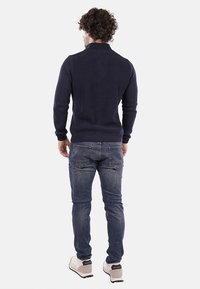 Maglione a coste blu navy con colletto alto, design aderente, indossato con jeans grigi e scarpe chiare. Vista da dietro.