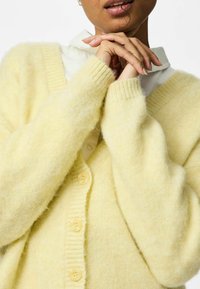 Gul fluffig kofta med ribbade muddar och femknappslås, lager-på-lager över en vit skjorta med krage. Mjuk textur, mysig design.