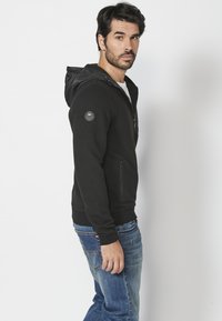 Felpa nera con zip e texture a costine, cappuccio regolabile, tasche frontali e toppa con logo sulla manica, abbinata a jeans blu.