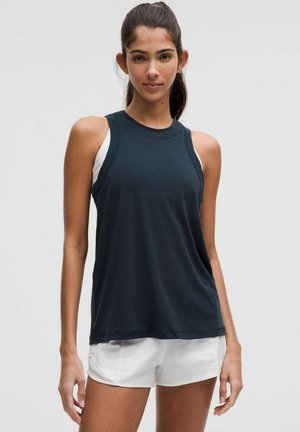 Mouwloos sporttopje van donkergrijze stof met een ronde halslijn en witte accenten. Draagt witte korte broek. Glad textuur.