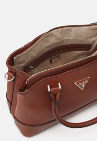 Bruine handtas met textuur, voorzien van dubbele handgrepen, gouden hardware en een open interieur met één ritsvak en het merkembleem.