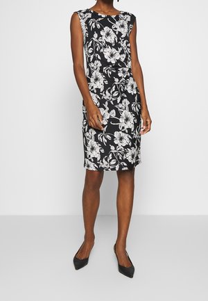 Vrouw draagt een mouwloze, knielange zwarte jurk met witte bloemenprint en zwarte schoenen met spitse neus, staand tegen een effen achtergrond.